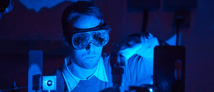 A man staring into a bright blue laser in the dark, wearing laser safety goggles